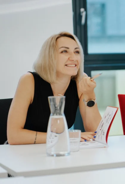 Eine Frau mit blondem Bob und ein glatzköpfiger Mann mit Bart sitzen einer Dritten Person gegenüber. Auf dem Tisch zwischen den Personen befinden sich ein Wasserkrug sowie ein Notizbuch.